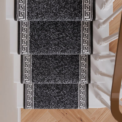 Four black and gray stair runner rugs with white Greek key borders, shown installed on a staircase with light wood flooring and white banister posts.