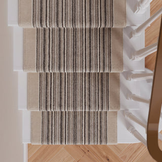 A set of beige stair runner rugs with vertical black and gray stripes, shown installed on a staircase with light wood flooring and white banister posts.
