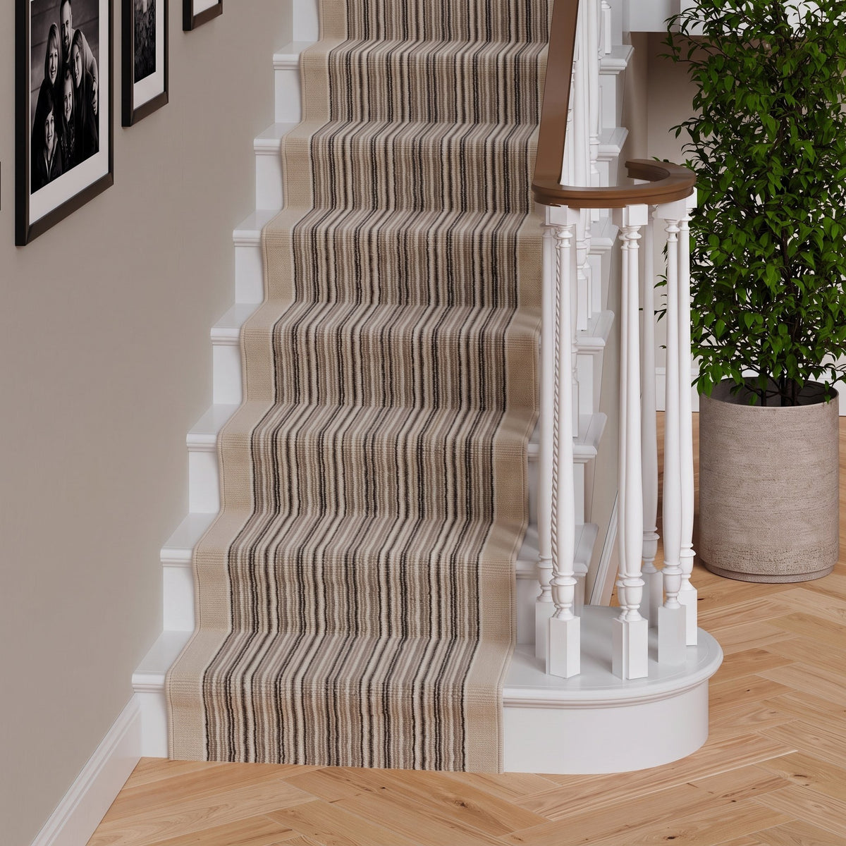 A beige and brown striped carpet runner installed on a white staircase in a home interior, with a wooden handrail, white balusters, framed wall art, and a potted green plant.
