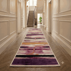 A long runner rug with an abstract purple, pink, and orange pattern is displayed on a herringbone wood floor in a classic, elegant hallway with cream paneled walls and a staircase in the background.