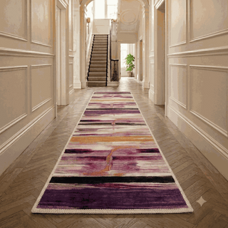 A long runner rug with an abstract purple, pink, and orange pattern is displayed on a herringbone wood floor in a classic, elegant hallway with cream paneled walls and a staircase in the background.