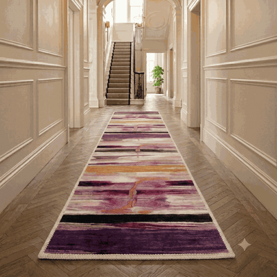A long runner rug with an abstract purple, pink, and orange pattern is displayed on a herringbone wood floor in a classic, elegant hallway with cream paneled walls and a staircase in the background.