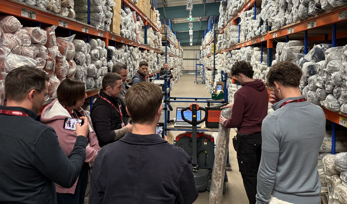 A group of people standing in a warehouse aisle surrounded by tall shelves packed with packaged textiles or fabrics, with one person holding a large rolled material.