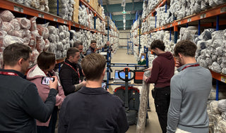 A group of people standing in a warehouse aisle surrounded by tall shelves packed with packaged textiles or fabrics, with one person holding a large rolled material.