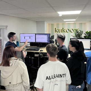 Group of people gathered around a desk with computer monitors, in an office setting with 'kukoon' branding.