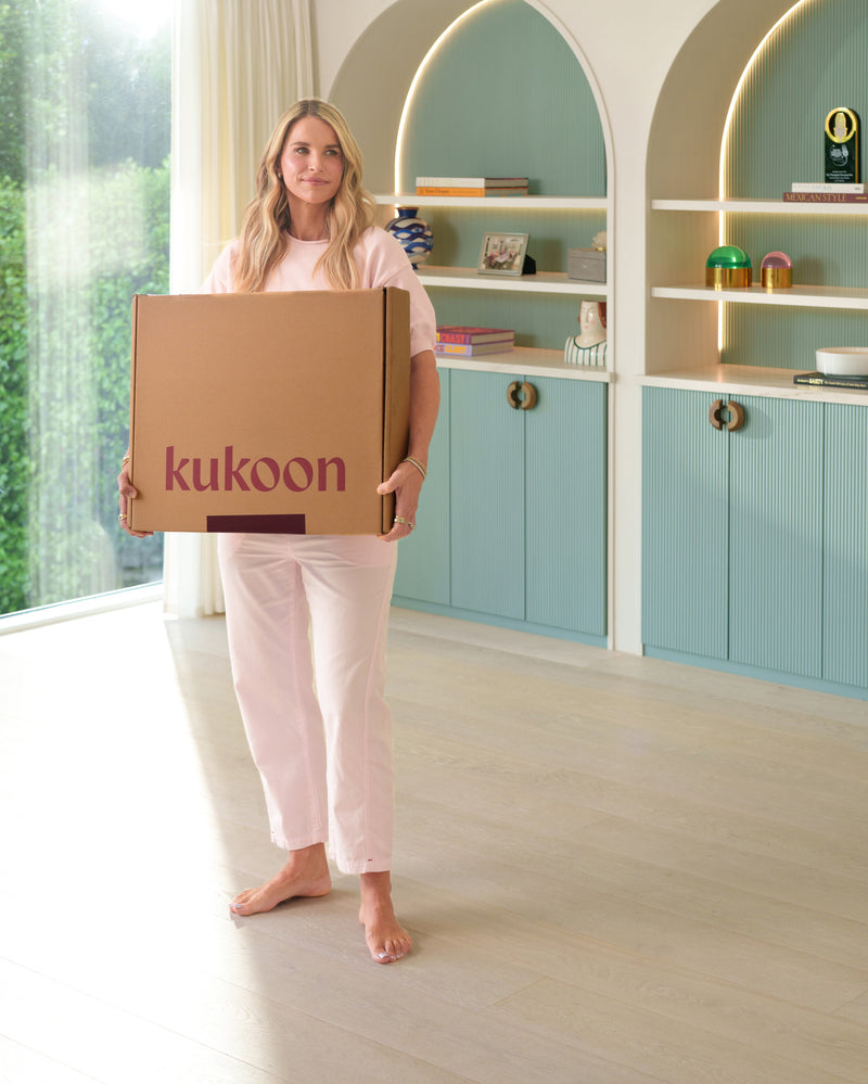 A woman in a pink outfit stands barefoot in a bright room holding a large brown cardboard box with the brand name 'kukoon' printed on the front. She is positioned in front of built-in shelves and cabinets in a pastel teal and cream color scheme.