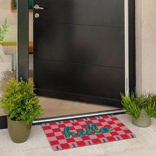 Black door with a red checkered doormat featuring the word 'hello', flanked by potted plants.
