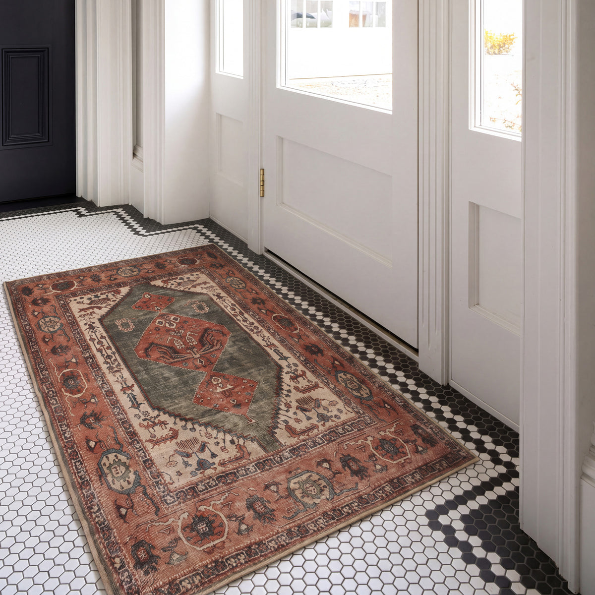 A rectangular vintage-style area rug with a distressed tribal pattern in rust red, beige, and gray, placed on a black and white hexagonal tile floor in front of white paneled doors.