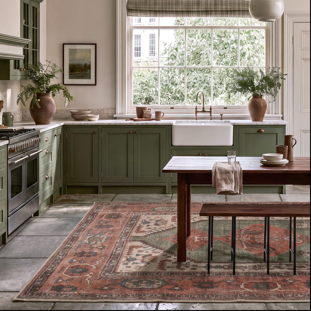 A styled kitchen interior with sage green cabinets, a large patterned area rug, a wooden dining table, and a window looking out onto green trees.