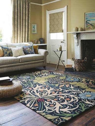 A large William Morris style floral area rug in navy, green, beige, and yellow is shown in a styled living room with a sofa, fireplace, and wood floors.