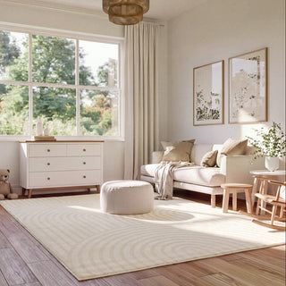 A large cream-colored area rug with a concentric wave pattern in a bright, modern living room. The room features a cream sofa, a matching ottoman, a white dresser, and light wood flooring.