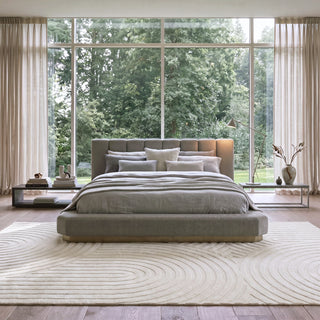A modern upholstered bed with a low platform frame and padded headboard, dressed in neutral gray bedding, set in a bright bedroom with large windows overlooking green trees. A light beige patterned rug covers the floor, with matching curtains and side tables on both sides.