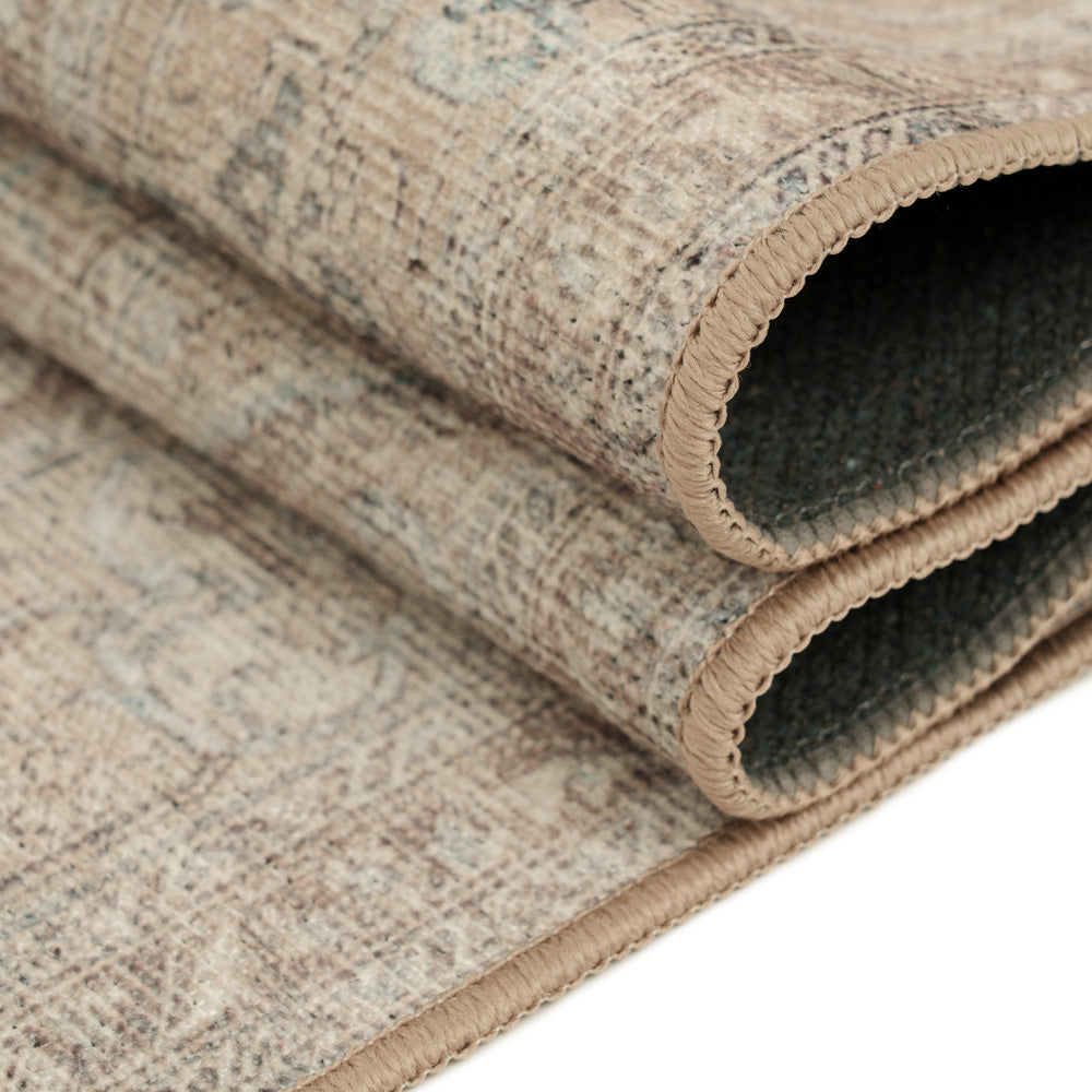 A close-up of a rolled area rug showing its distressed beige and brown patterned top and a dark charcoal gray underside, with a tan stitched border.