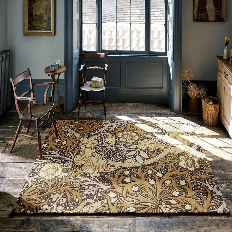 A large rectangular area rug with an intricate botanical and floral pattern in beige, brown, and cream tones, displayed on a rustic wooden floor in a styled room with vintage wooden chairs and a window.