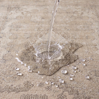 A close-up of a beige patterned rug with water being poured onto its surface, showing strong water repellency and a small splash.
