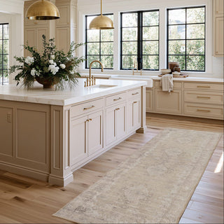 A long beige runner rug with a subtle distressed pattern is styled on a light wood floor in a bright, modern kitchen with cream cabinets, white countertops, and gold pendant lights.