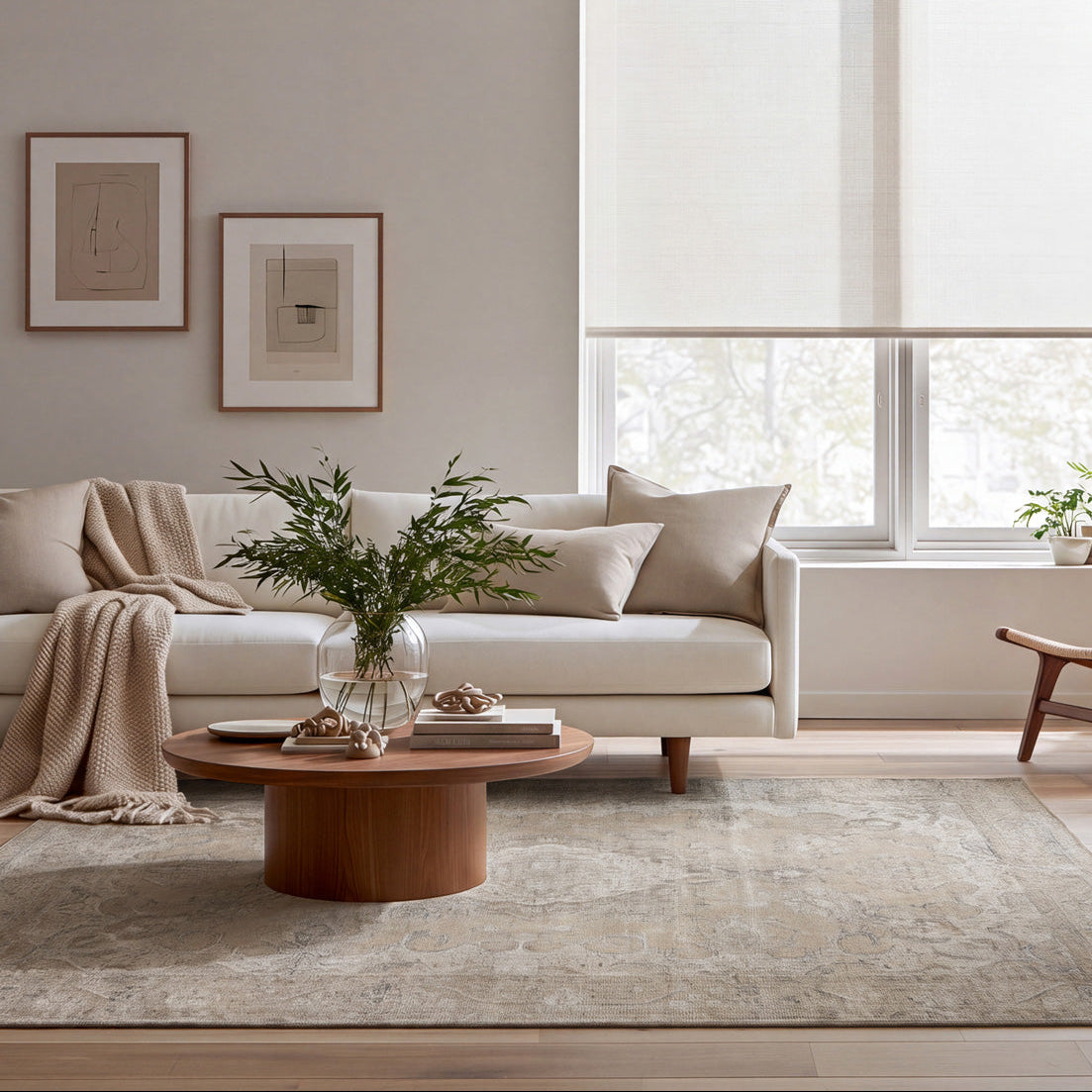 A modern living room with a light beige sofa, a round wooden coffee table, a textured area rug, and a large window with off-white roller blinds. The room is styled with neutral-toned furniture, framed wall art, and potted plants.