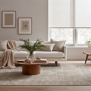 A modern living room with a light beige sofa, a round wooden coffee table, a textured area rug, and a large window with off-white roller blinds. The room is styled with neutral-toned furniture, framed wall art, and potted plants.